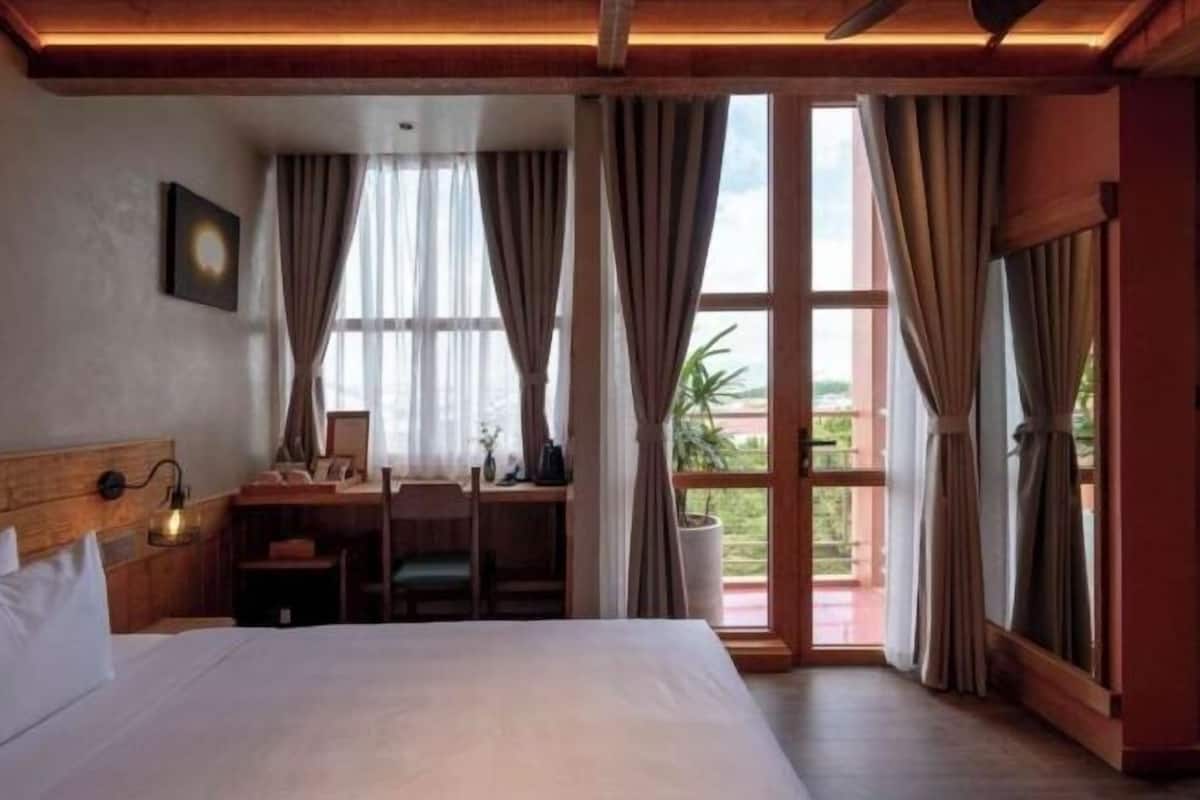 A hotel room with a large bed, a desk with a chair, a nightstand, and a window with curtains.