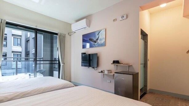 A hotel room with two beds, a television, a small refrigerator, and a window with a view of buildings. - Dolphin B&B (Pingtung)