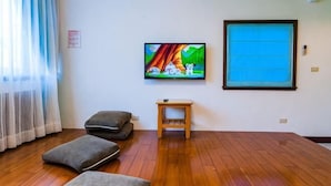 Ein Zimmer mit Holzboden, einem an der Wand montierten Fernseher, einem kleinen Holztisch und einem Fenster mit blauer Jalousie.