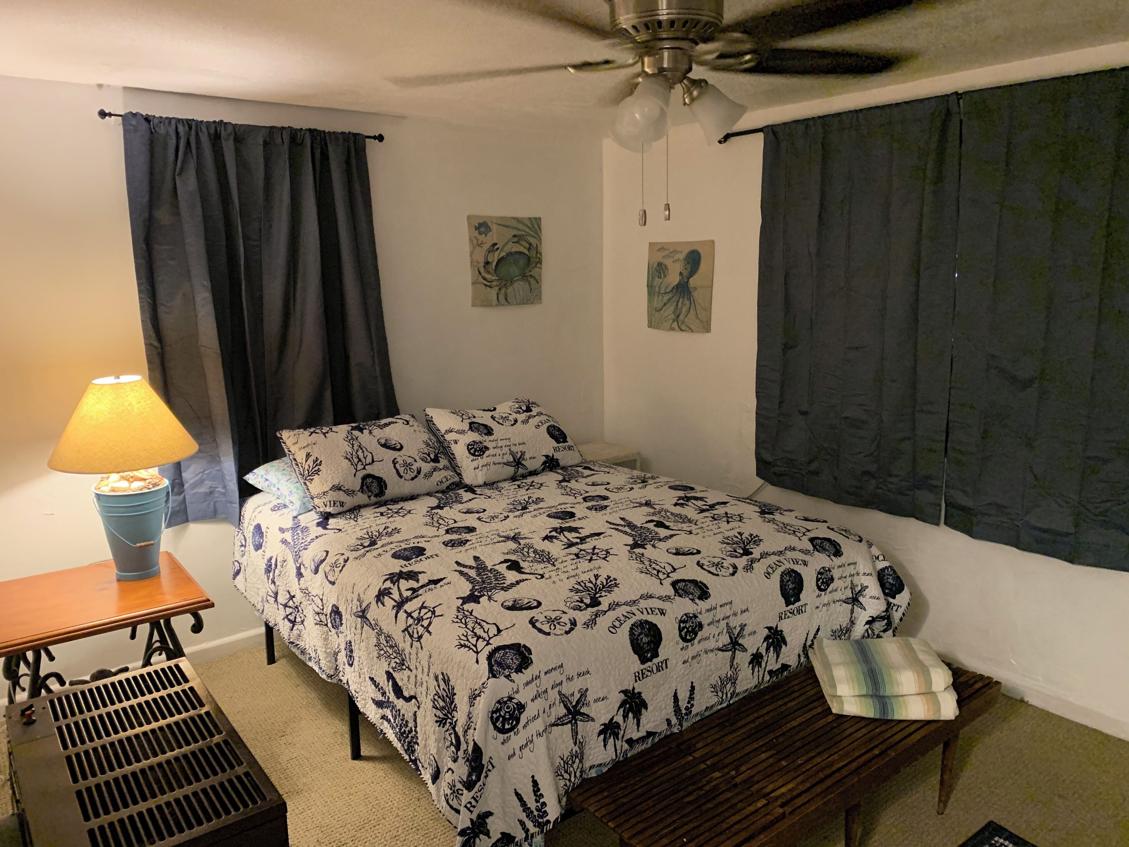 A bedroom with a bed, a desk with a lamp, a ceiling fan, and a window with curtains.