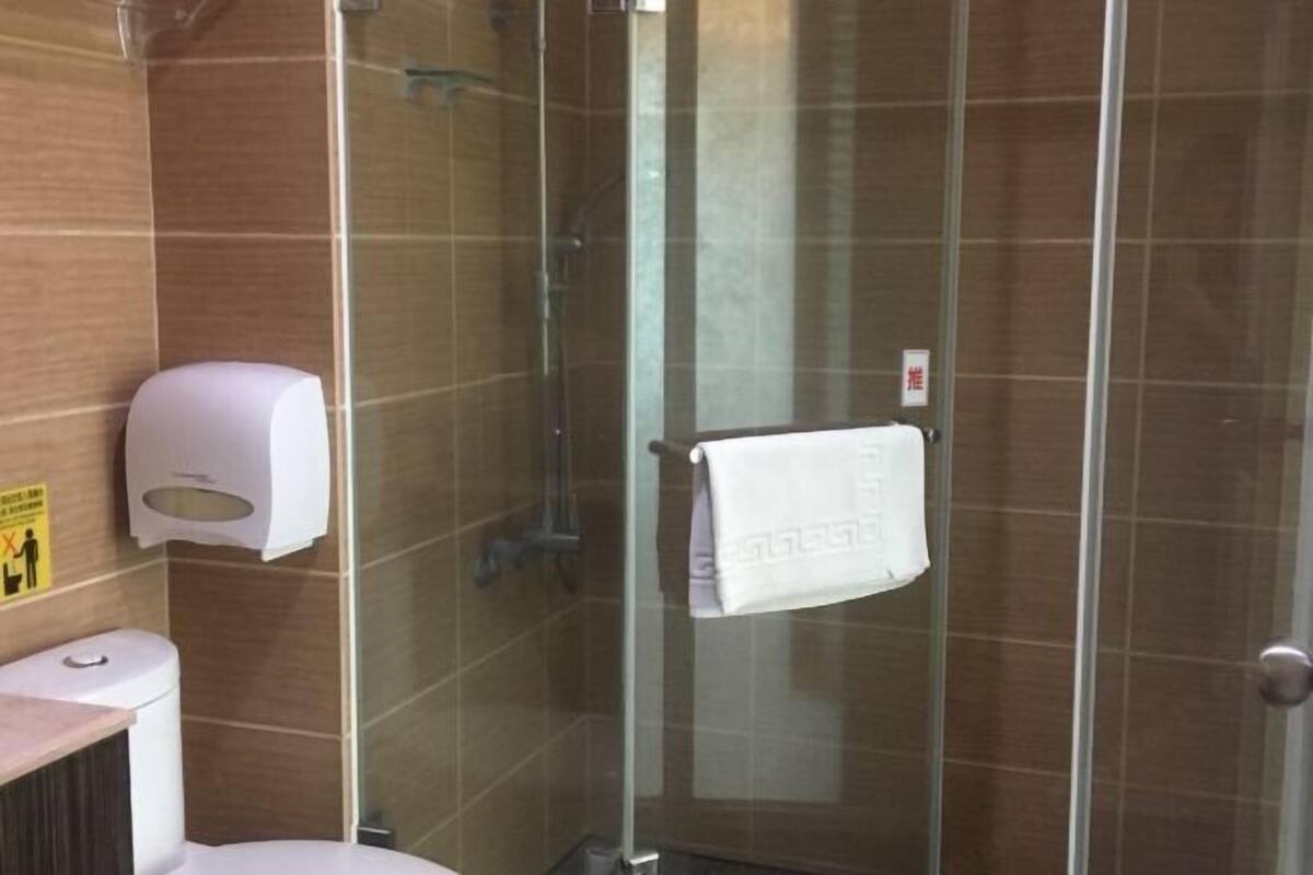Un box de ducha con paredes de cristal, una toalla blanca colgada en un perchero, un dispensador de gel hidroalcohólico en la pared y un inodoro con separador.