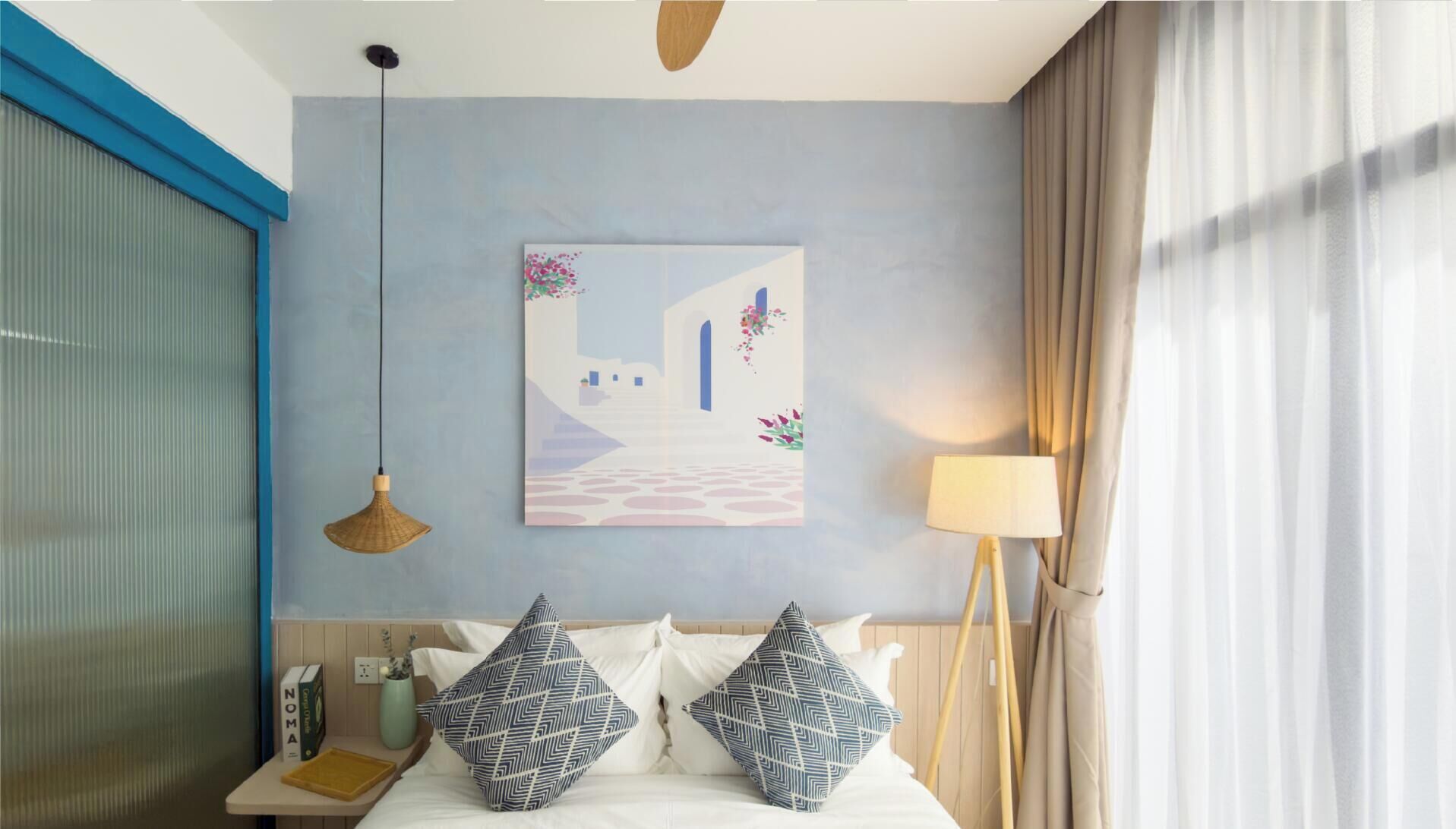 A hotel room with a bed, a bedside table with a book and a vase, a wall-mounted artwork, a hanging lamp, and a window with curtains.