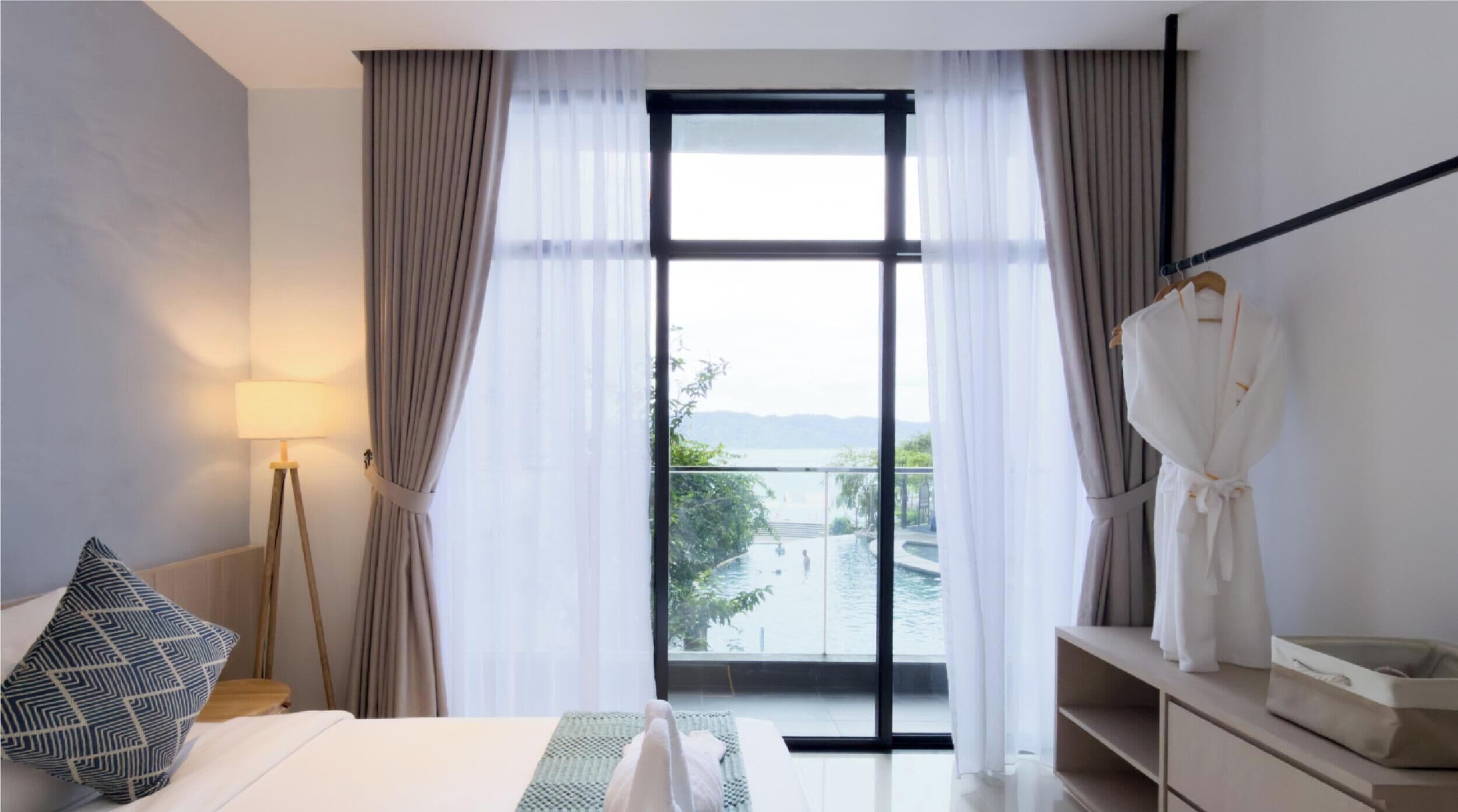 Ett hotellrum med en säng, en sänglampa, en badrock som hänger på en krok och utsikt över en pool och berg genom ett stort fönster.