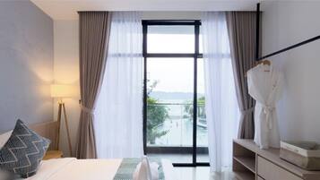 Une chambre d’hôtel avec un lit, une lampe de chevet, une robe suspendue et une vue sur une piscine et des montagnes, le tout visible à travers une grande fenêtre.