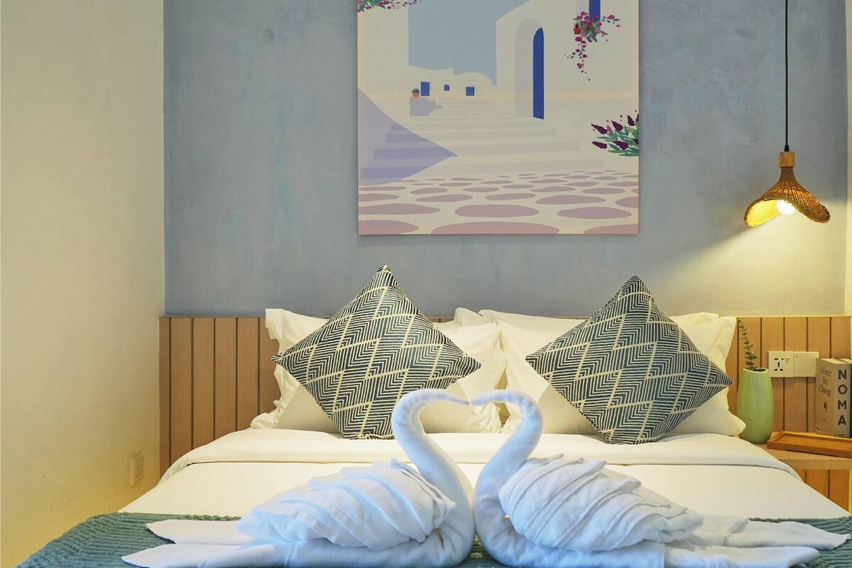 A hotel room with a bed, two patterned pillows, a wall-mounted painting, and a hanging light fixture.