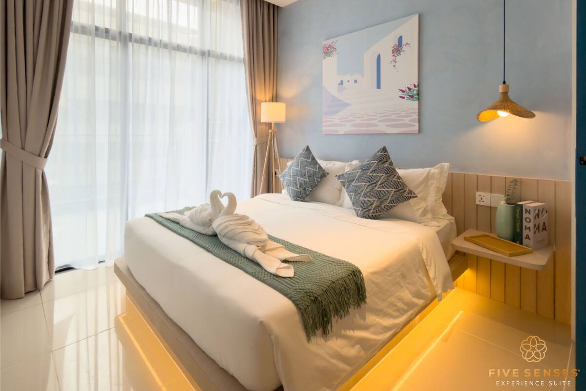 A neatly made bed with a green throw blanket and a white towel, a bedside table with a lamp, a painting on the wall, and a hanging light fixture.