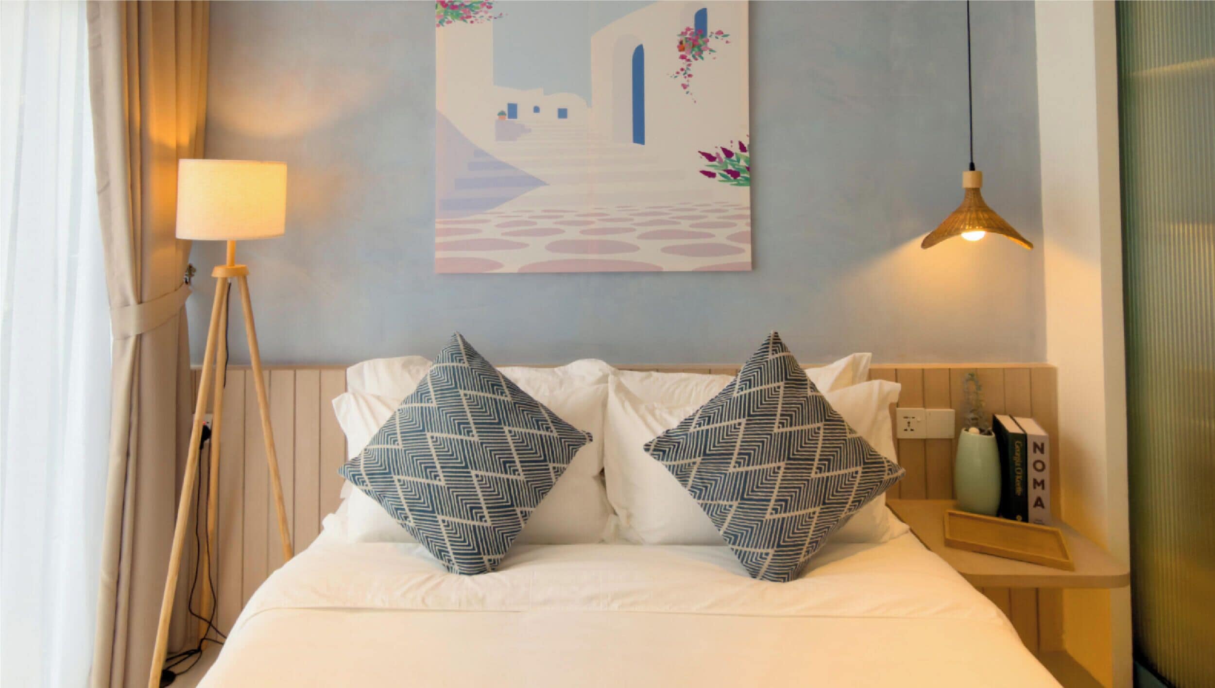 Un lit bien fait, avec du linge de lit blanc et deux coussins à motifs, une lampe de chevet, une œuvre d’art accrochée au mur représentant une scène de rue et une suspension lumineuse.