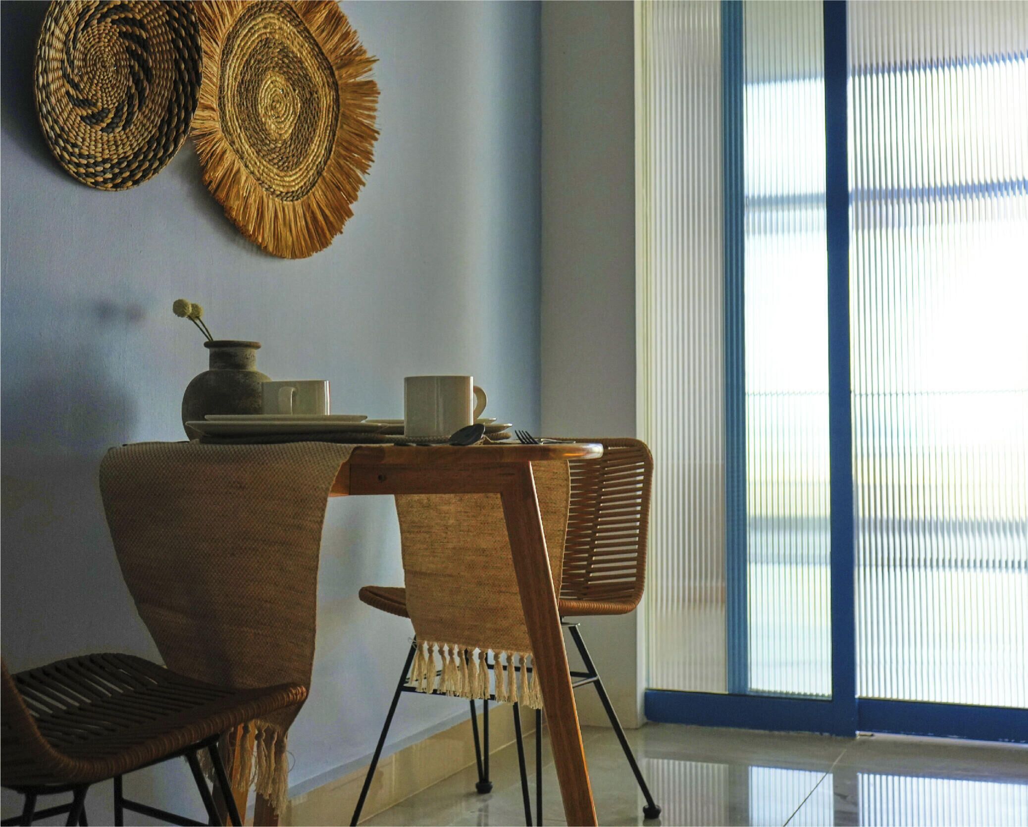 A dining area with a table set for two, two woven wall hangings, and a window with blinds.