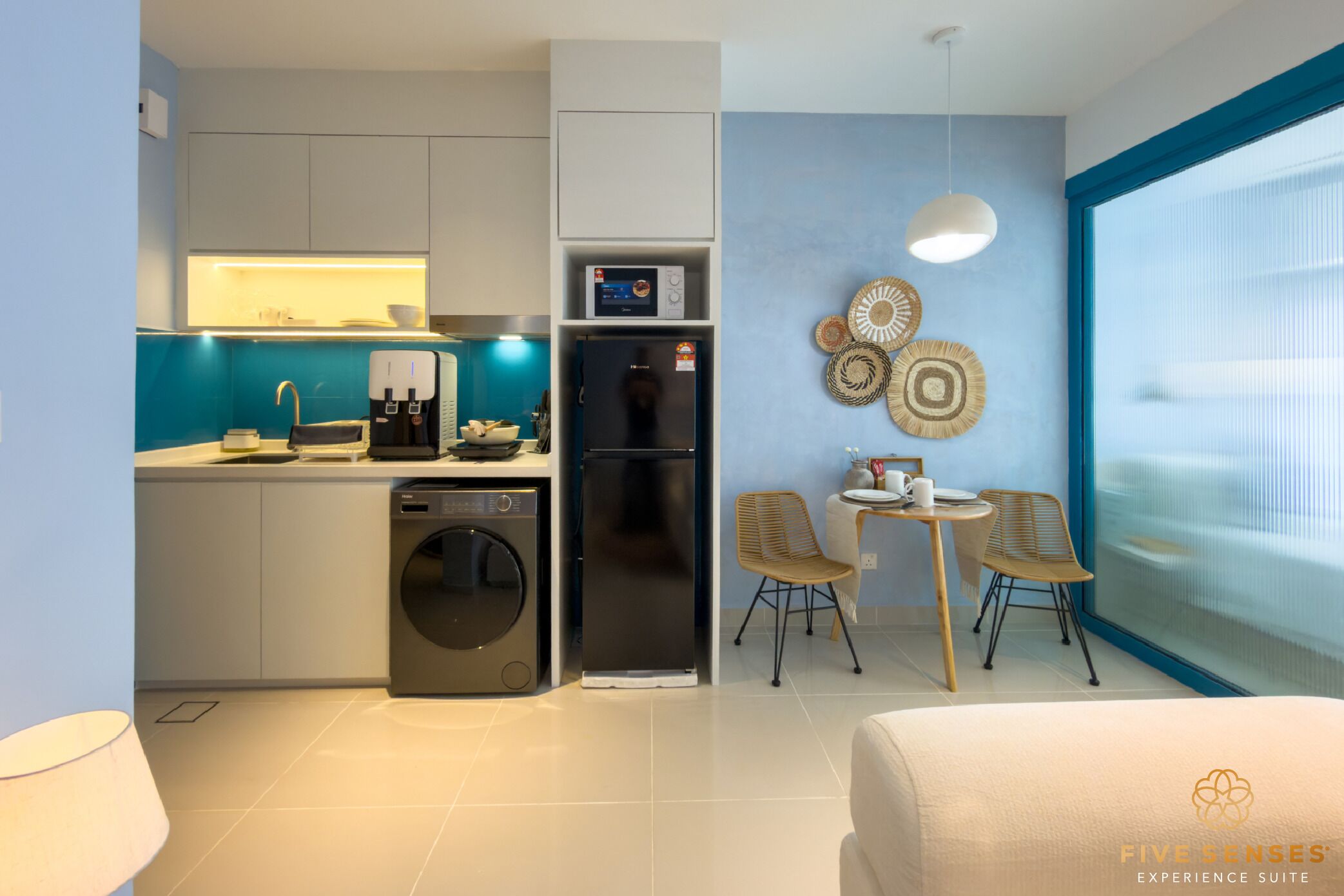 A modern kitchen with a dishwasher, washing machine, and a coffee machine. There is a dining area with two chairs and a table. The room has a large window and decorative wall art.