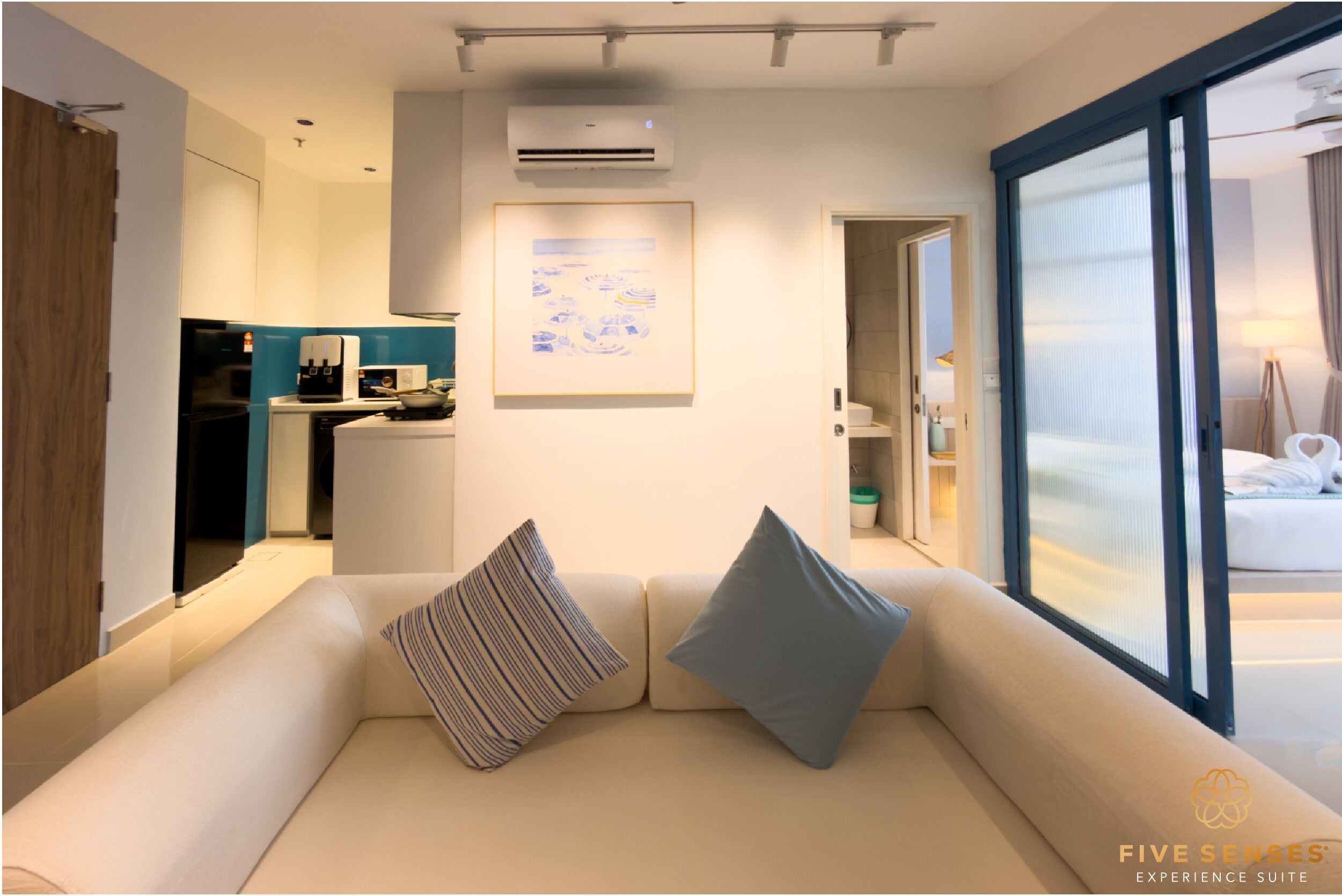 A modern hotel room with a sofa, a kitchen area, and a large window.