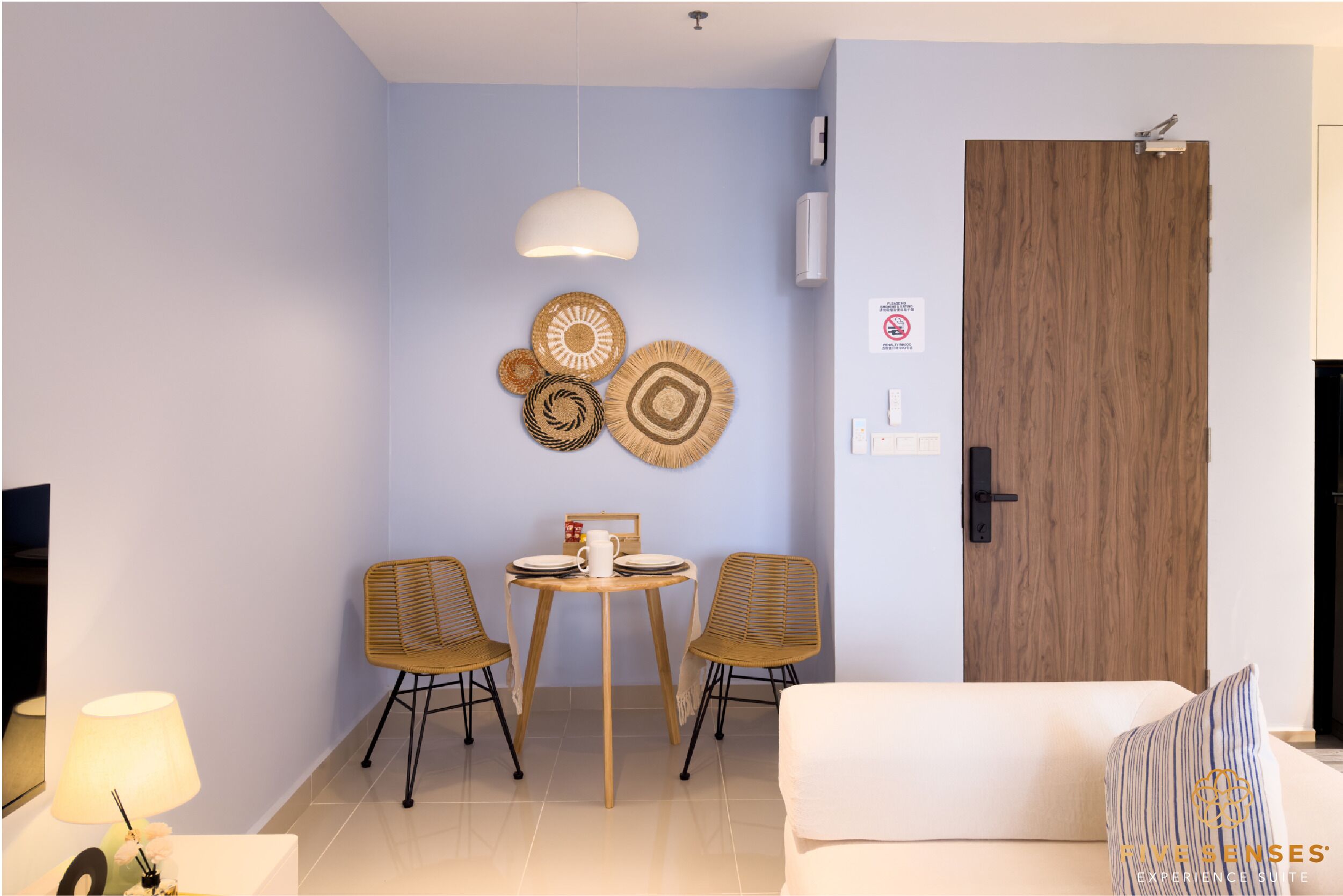 A modern living room with a sofa, two wicker chairs, a dining table with plates and a mug, a hanging light fixture, and a wooden door.