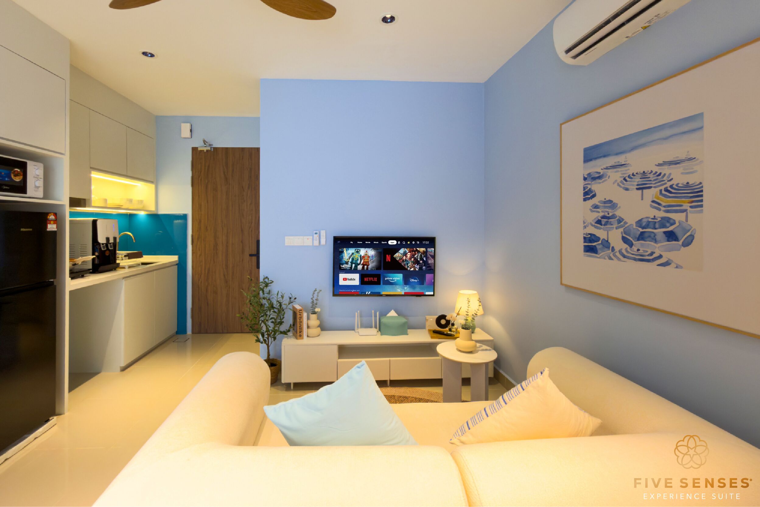 Een moderne woonkamer met een bank, een aan de muur bevestigde televisie, en een keuken met een koelkast en een magnetron.