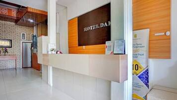 Ein Hotelrezeptionstresen mit Holzbeschlag und einem Schild mit der Aufschrift "HOTEL DAK". An der Wand hängt ein gerahmtes Bild, und ein Schwarzes Brett wirbt mit einem Rabatt von 10%.