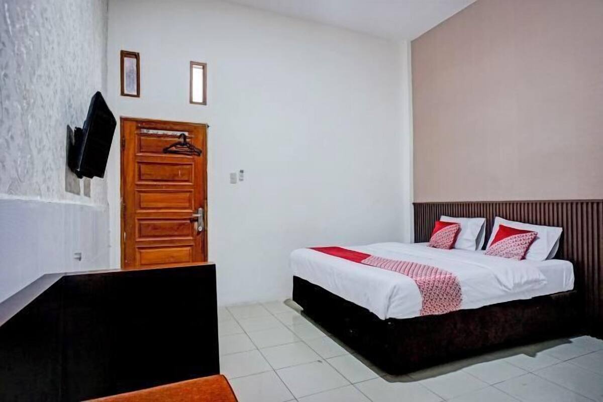 Une chambre à coucher avec un lit, une porte en bois, un téléviseur fixé au mur et une petite fenêtre.