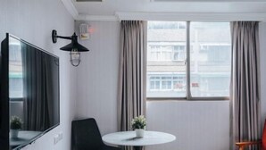 A hotel room with a flat-screen TV, a round table with a potted plant, a black chair, a wooden chair, and a window with curtains. - Meworld Hotel-Sanchong (New Taipei City)