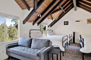 Ein Schlafzimmer mit zwei Betten, einem Schlafsofa und Blick auf Bäume durch große Fenster.