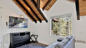 A modern living room with a grey sofa, a flat-screen TV mounted on the wall, a wooden ceiling, and a large window with a view of trees. - Round Rock Lodge at Big Bear | Boulder Bay Modern Retreat w/ Pool Table | Minutes to Activities (Big Bear Lake)
