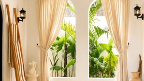A spa room with a hot tub, two windows with curtains, a wooden chair, a small table, and a wall-mounted lamp. - Okeanos Boutique Hotel Ungasan Uluwatu (Ungasan)
