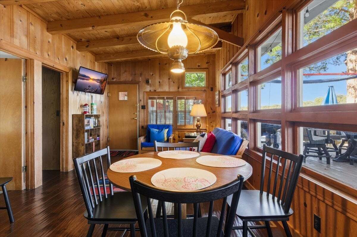 A wooden interior with a round table, chairs, a television, and large windows.