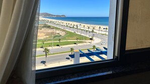 Blick aus einem Fenster auf einen Strand mit Palmen, eine Straße und ein ferner Berg.