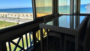 Ein Balkon mit Glastisch und Korbstühlen, mit Blick auf einen Strand mit Palmen und wolkenlosem, blauem Himmel.