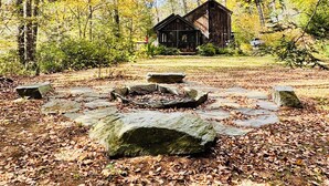 Una cabaña rústica rodeada de un bosque con hojas otoñales, un sendero de piedra y un cielo despejado.