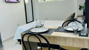 Un comedor moderno con una mesa de madera, dos sillas negras con asientos de mimbre, un televisor en la pared y una ventana con vista a la vegetación.