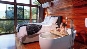 A bedroom with a large bed, a bathtub, and a view of trees through large windows. - Recanto do Bicí (Urubici)