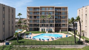 Een meerlaags hotel met een zwembad, omringd door ligbedden en palmbomen.