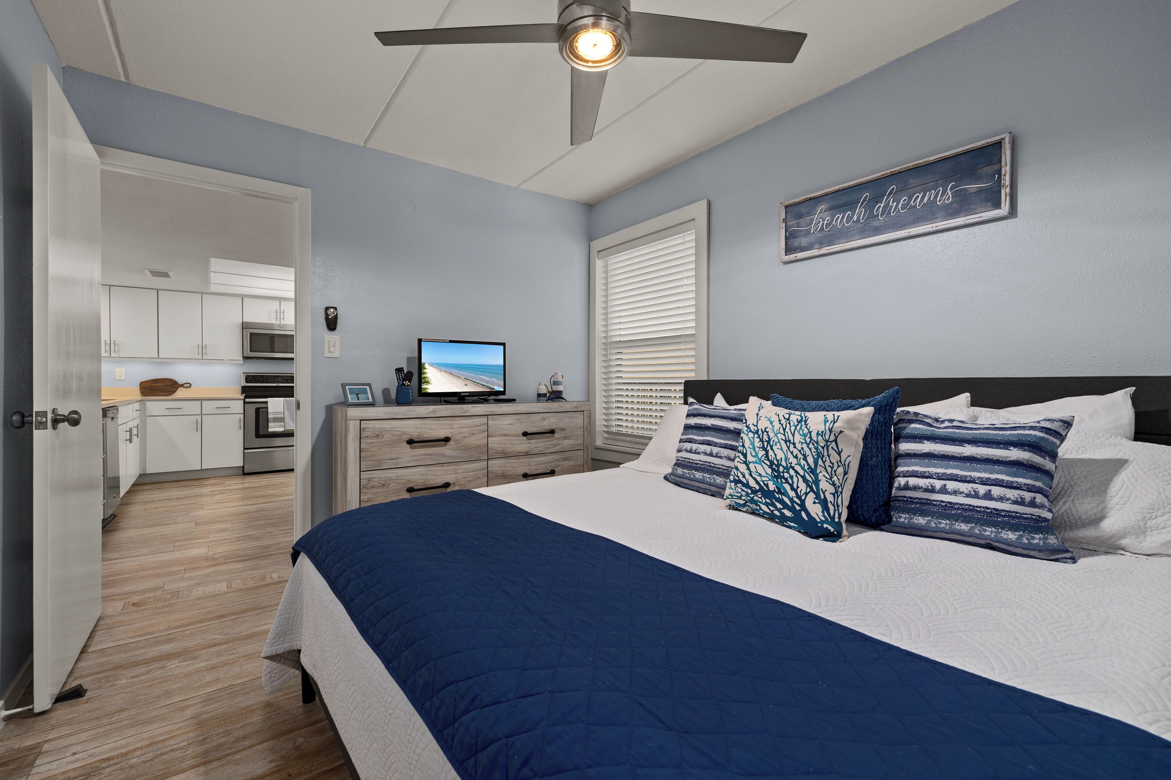 A bedroom with a bed, a ceiling fan, a TV, and a kitchen in the background.