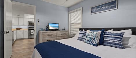 A bedroom with a bed, a ceiling fan, a TV, and a kitchen in the background.