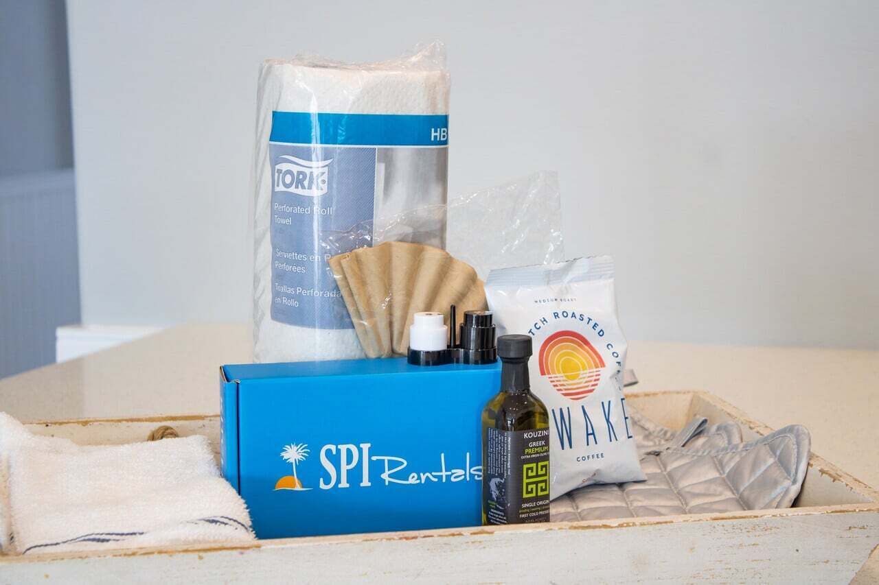 A hotel room with a blue box labeled 'SPI Rentals,' a bag of 'HBI' towels, a 'WANK' coffee bag, a bottle of 'Kouzini Greek Premium Coffee,' and a fan.