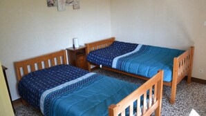 Huoneessa on kaksi puista sänkyä, pieni yöpöytä, jossa on lamppu, ja seinä, jossa on kehystettyjä kuvia.