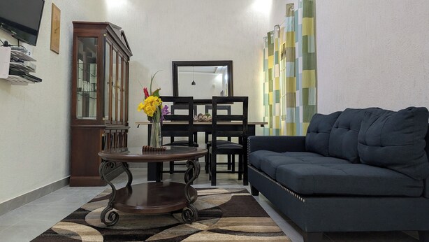 Un salón con un sofá azul oscuro, un armario de madera, una mesita con un jarrón de flores y una alfombra estampada.