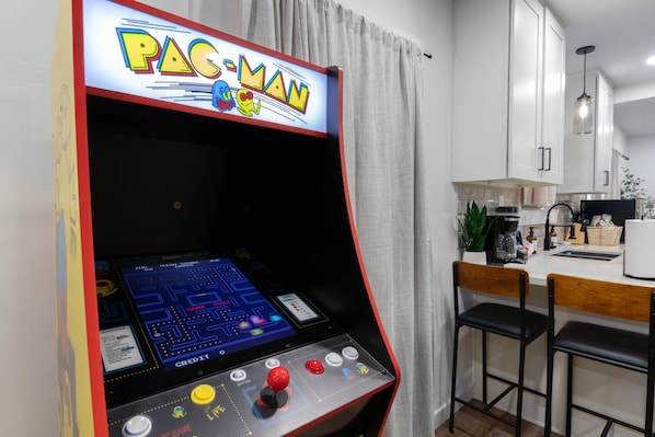 Une borne d’arcade Pac-Man dans une cuisine moderne avec des meubles blancs, une machine à café et un coin repas avec deux chaises.