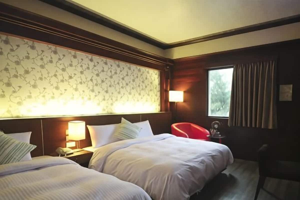 A hotel room with two beds, a red chair, a window with curtains, and a wall with floral wallpaper.