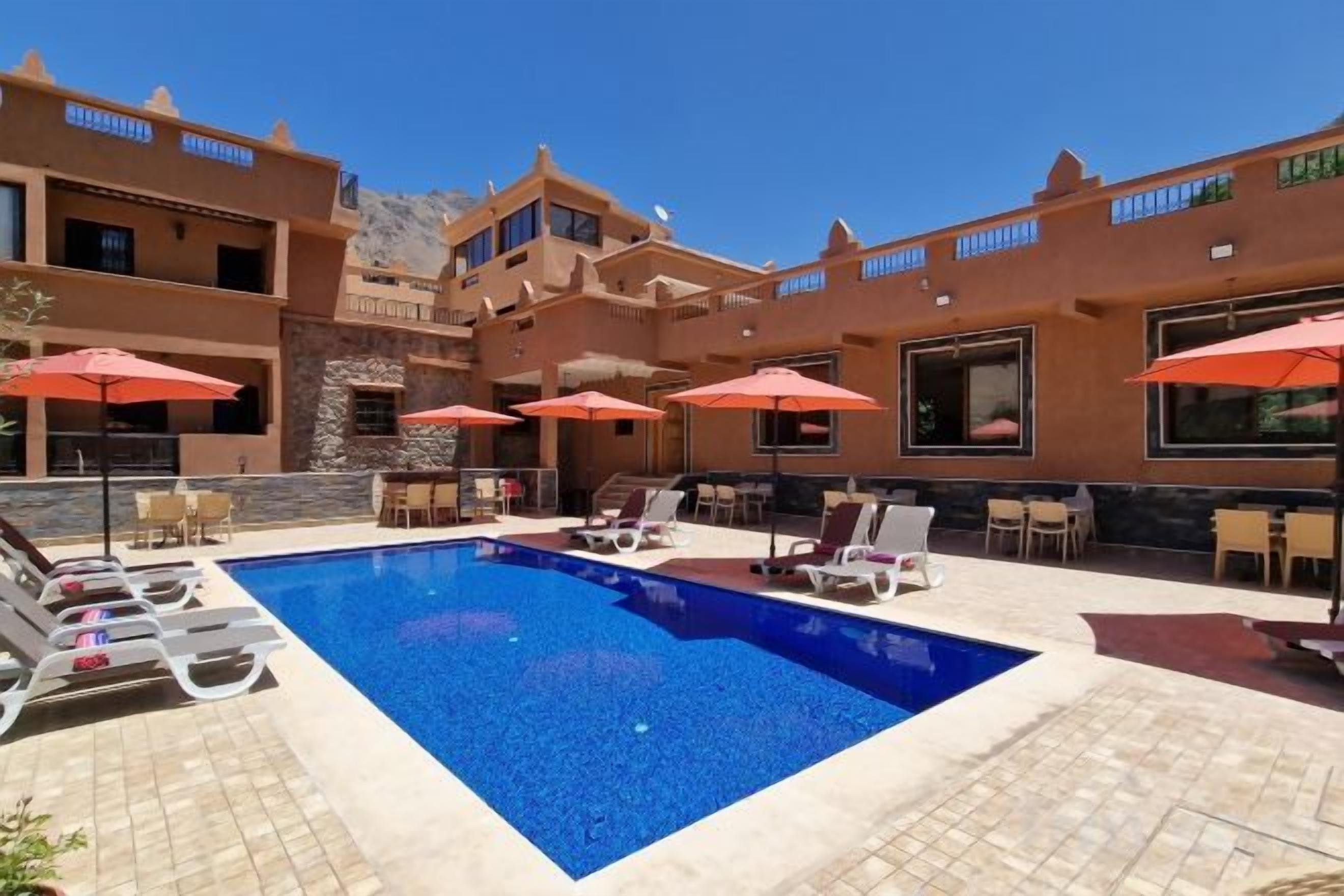 Zona piscina con sedie a sdraio e ombrelloni, circondata da un edificio con numerosi balconi.