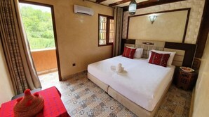 Ein Schlafzimmer mit einem großen Bett, einem kleinen Tisch und einem dekorativen Gegenstand, einem Fenster mit Blick auf Grünflächen und einem Deckenventilator.