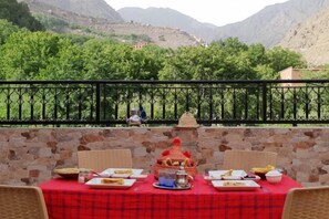 Una mesa lista para una comida, con mantel rojo, rodeada de muros de piedra, y vista a árboles verdes y montañas al fondo.