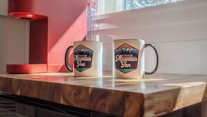 Twee mokken met een bergdesign op een houten oppervlak, naast een rode koffiezetapparaat.
