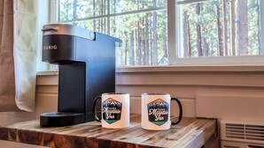 Een koffiezetapparaat en twee mokken op een houten tafel bij een raam met uitzicht op een bos.