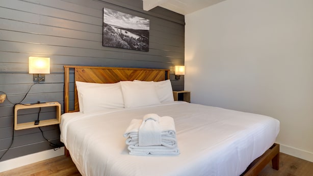A neatly made bed with white linens and a folded towel, wooden headboard, bedside lamps, and a framed picture on the wall. - Tahoe Mountain Inn #34 (SOUTH LAKE TAHOE)