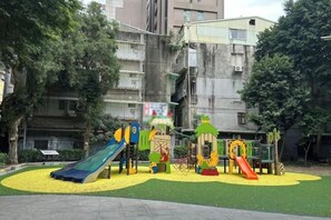 A playground with slides and climbing structures, surrounded by a green area and residential buildings in the background. - Meworld Hotel Zhongshan (Taipei)