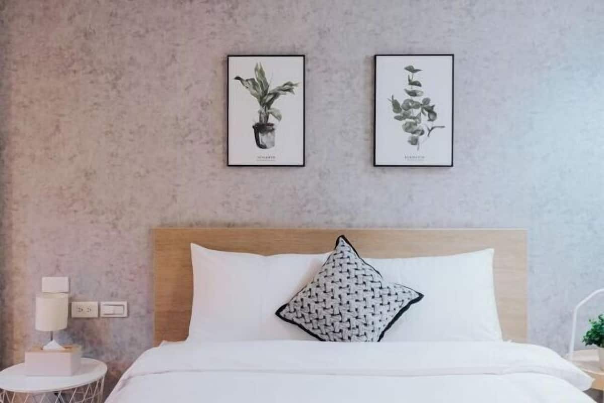 A bedroom with a wooden headboard, white bedding, a round bedside table with a lamp, and two framed botanical prints on the wall.