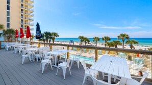 Une terrasse avec des tables et des chaises blanches, une piscine et une vue sur la plage et l’océan.