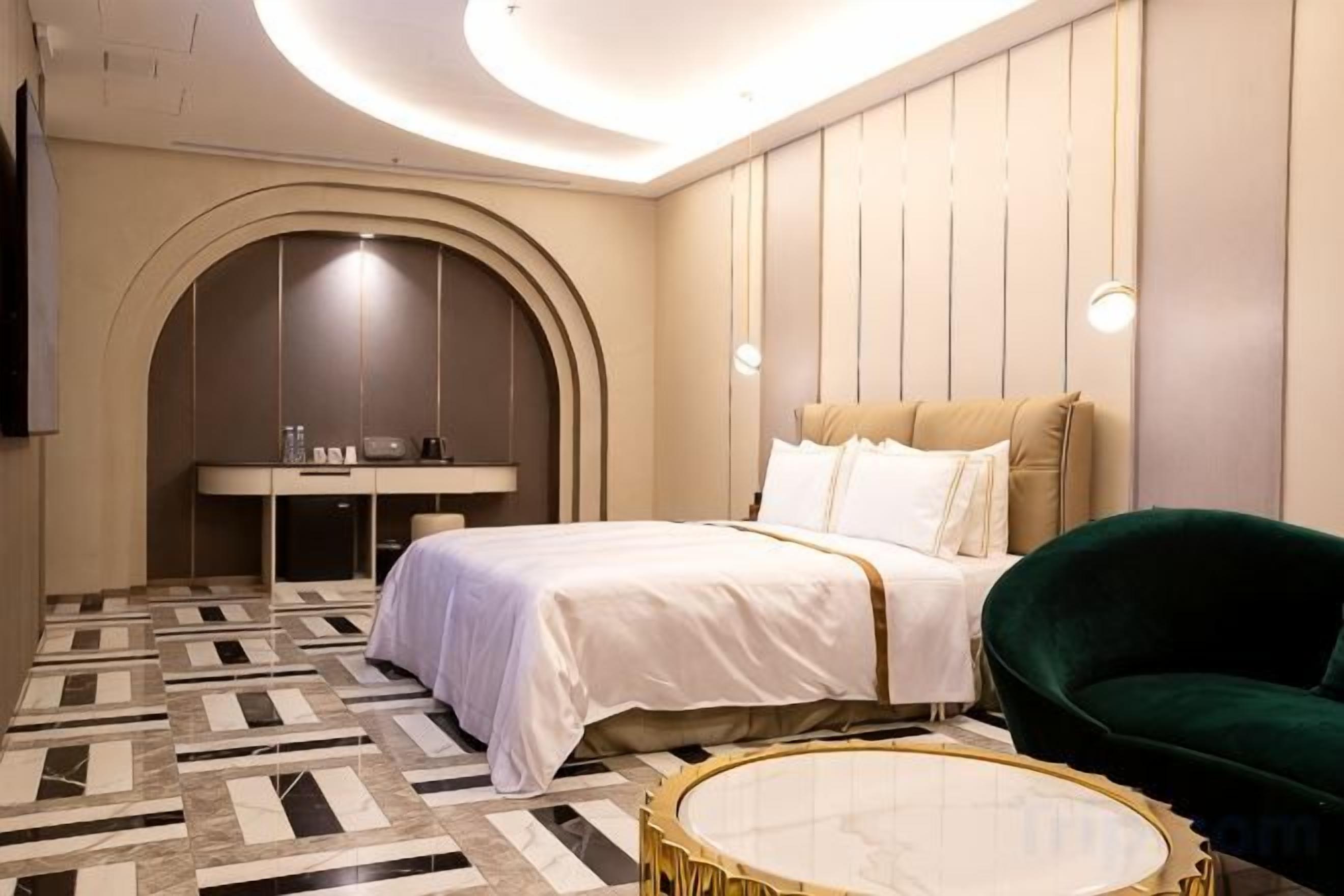 Una habitación de hotel con una cama, una mesa redonda, un sillón verde, un escritorio y un televisor.