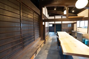Una habitación amplia con una larga mesa de madera y bancos, una zona de cocina con electrodomésticos modernos, y un techo alto con vigas a la vista.