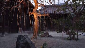 Un jardín japonés tradicional con una gran roca, un árbol de ramas colgantes y un edificio al fondo.