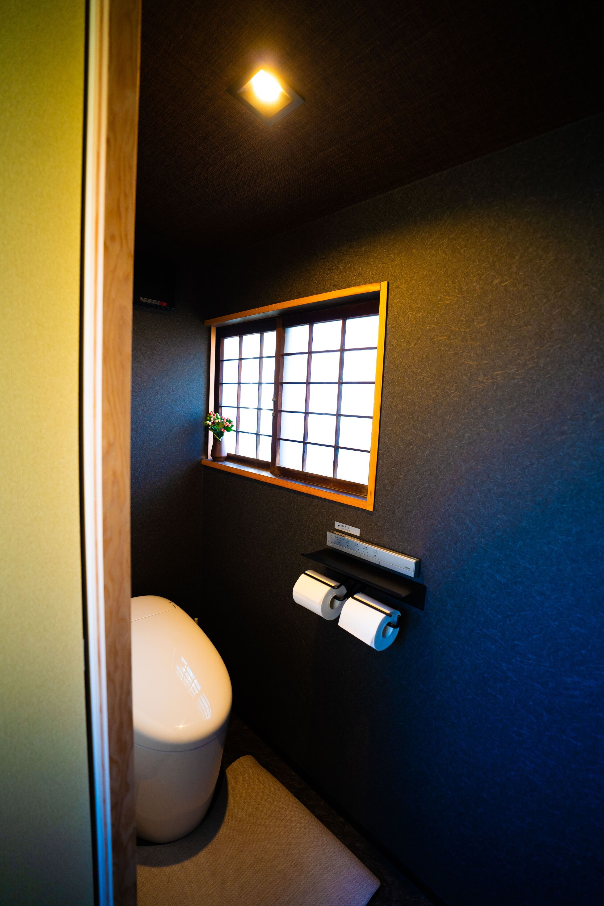 A small bathroom with a toilet, a window, and a shelf with a roll of toilet paper.