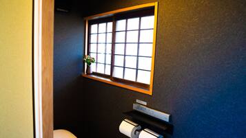 A small bathroom with a toilet, a window, and a shelf with a roll of toilet paper.